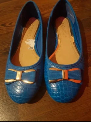 Blå ballerinaskor med rosett - Snygga blå ballerinaskor med krokodilpräglat mönster och dekorativ rosett framtill med guldfärgade detaljer. Köptes på Vinted, storlek 38 men små för mig så gissar att den passar mer 37 eller 37,5