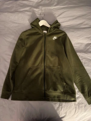 Olivgrön zip hoodie från Nike - Snygg olivgrön hoodie från Nike med dragkedja framtill och vit logga på bröstet. Hoodien har huva, två stora fickor fram och är tillverkad i mjukt sweatshirtmaterial. Perfekt för en avslappnade och sportig stil. Storlek XL i barn så sitter som S i vuxen
