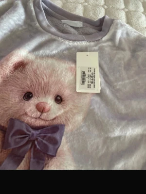 Ljuslila loungewear med nalletryck - Supersöt ljuslila loungewear-tröja i mjukt, fluffigt material. Framsidan har ett stort tryck av en rosa nallebjörn med lila rosett. Perfekt för mysiga hemmakvällar och chill dagar. Rund halsringning och avslappnad passform.
