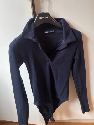 Mörkblå långärmad body med krage - Snygg mörkblå body från Zara med lång ärm och klassisk krage. Modellen har djup v-ringning och är tillverkad i ett mjukt, stretchigt material som sitter skönt mot kroppen. Perfekt att styla med jeans eller kjol för en trendig look.