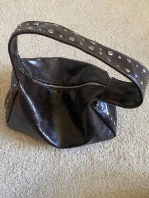 Gina Svart axelväska i skinnimitation - Cool svart handväska i skinnimitation med bred axelrem dekorerad med stora silverfärgade nitar. Väskan har en mjuk, slouchy form och stängs med dragkedja upptill. Perfekt för dig som gillar en edgy och trendig look.