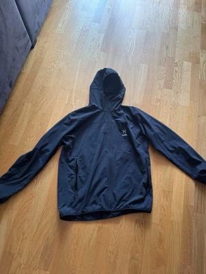 Svart vindjacka med huva Haglöfs - Snygg svart vindjacka från Haglöfs med huva och dragkedja framtill. Två sidofickor med dragkedja och diskret logga på bröstet. Lätt material som passar perfekt för blåsiga dagar. Enkel och stilren design.