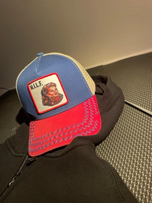 M.I.L.F. Keps från Leo LIMITED - Kepsen är limited då den har gjort om företaget från kepsar och Annat till parfymer Säljer en unik truckerkeps med blå framsida, röd skärm och beige mesh baktill. Kepsen har en stor patch framtill med texten 'M.I.L.F.' och en retroillustration av en kvinna. Skärmen har blå sicksacksömmar som kontrast. Perfekt statement-piece för dig som vill sticka ut.