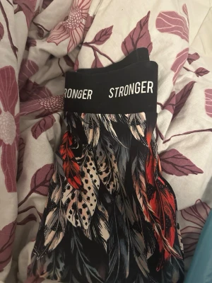 Stronger leggings med fjädermönster - Snygga leggings från Stronger med bred svart midjeresår och logga. Mönstret består av färgglada fjädrar i svart, rött, beige och grått. Perfekta för träning eller chill. Materialet är stretchigt och följsamt för bästa komfort.