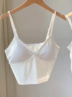 Vit ribbad body med spetsdetaljer - Säljer en vit ribbad body med tunna axelband och fina spetsdetaljer längs urringningen. Materialet är mjukt och stretchigt, perfekt för en bekväm och snygg look. Passar perfekt under tröjor eller som ett statement-plagg.