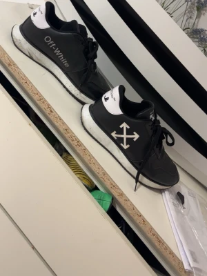 Svarta Off-White sneakers med pilar - Snygga svarta sneakers från Off-White med vita detaljer och den ikoniska pil-loggan på sidan. Skorna har en vit sula med svarta prickar och svart snörning. Materialet ser ut att vara en mix av läder och textil. Perfekta för dig som gillar streetwear och vill sticka ut.