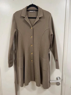 Beige lång kappa med knappar - Stilren beige kappa med klassisk krage och guldknappar framtill. Kappan har två stora fickor, markerad midja med bälte och långa ärmar. Materialet ser ut att vara mjukt och stickat, perfekt för lager på lager under kyliga dagar.