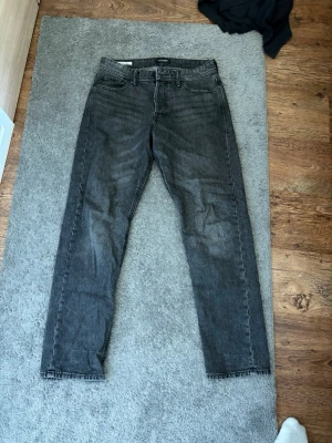 Svarta raka jeans med knappgylf - Säljer ett par svarta jeans med raka ben och knappgylf. Jeansen har klassisk femficksdesign och är tillverkade i denim med en tvättad look. Perfekta för en avslappnad och stilren outfit.