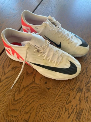 Inomhus skor Nike vita fotbollsskor  - Säljer ett par vita Nike fotbollsskor( inomhus)  med svart swoosh och neonrosa detaljer på hälen. Skorna har snörning och är tillverkade i syntetmaterial med låg profil och mönstrad ovansida för bra bollkontroll. Perfekta för dig som vill ha stil och funktion på planen. Sparsamt använda storlek 44,5 ord pris 1499kr