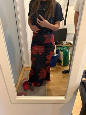 Långkjol med stora röda blommor - Säljer en långkjol med svart bas och stora röda blommor. Kjolen har en figurnära passform och är tillverkad i ett mjukt, stretchigt material som ger en snygg siluett. Perfekt för dig som vill sticka ut med ett färgstarkt mönster.