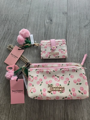 Rosa handväska med körsbär Juicy Couture - Söt handväska från Juicy Couture i ljusrosa med körsbärsmönster och guldfärgad logga. Väskan har matchande korthållare, guldkedja och fluffiga rosa nyckelringar. Perfekt accessoar för dig som gillar lekfull och trendig stil.