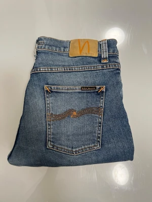 Blå jeans från Nudie Jeans - Snygga blå jeans från Nudie Jeans med klassisk femficksmodell och kontrastsömmar. Jeansen har en ljus tvätt och detaljer i orange, samt Nudie-logga på bakfickan och läderpatch i midjan. Perfekta för en avslappnad och trendig stil.