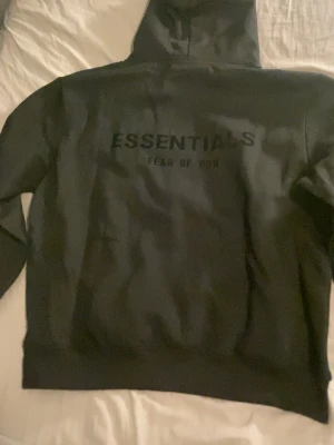 Svart Essentials Fear of God hoodie - Säljer en svart hoodie från Essentials Fear of God med tryck på både bröstet och ryggen. Klassisk huva och magficka framtill. Hoodien har en clean och stilren look, perfekt för dig som gillar streetwear och vill ha något som sticker ut utan att vara för mycket.