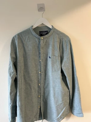 Grå långärmad henley från Abercrombie - Snygg blå långärmad henley-skjorta från Abercrombie & Fitch med rund halsringning och knappar framtill. Skjortan har en bröstficka med liten broderad logga och är tillverkad i mjuk bomull. Perfekt för en avslappnad stil.