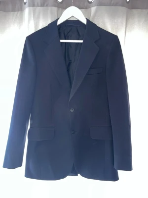Klassisk svart kavaj med två knappar - Stilren svart kavaj med två knappar framtill, klassiska slag och två fickor med lock. Kavajen har en rak passform och är helfodrad, perfekt för en dressad look. Materialet ser ut att vara av ullblandning för en snygg finish.