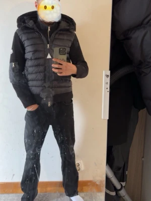 Svart pufferjacka med huva - Säljer en svart pufferjacka med huva och dragkedja. Jackan har quiltade partier på framsidan och ryggen samt stickade ärmar och sidor. Fickor med dragkedja och en patch på ärmen ger extra detaljerad look.