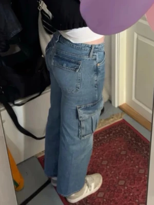 Blå cargobyxor i denim - Säljer ett par ljusblå cargobyxor i denim med raka ben och stora sidofickor. Klassisk femficksmodell med knapp och dragkedja framtill. Perfekta för en avslappnad och trendig look.