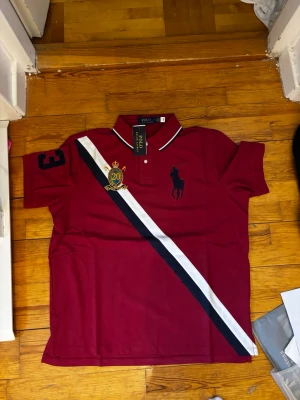 Vinröd pikétröja från Polo Ralph Lauren - Vinröd pikétröja med klassisk krage, broderad logga och emblem på bröstet samt kontrasterande vita och marinblå ränder. Kortärmad modell med nummerdetalj på ärmen och knappslå framtill. Perfekt för en sportig och stilren look.