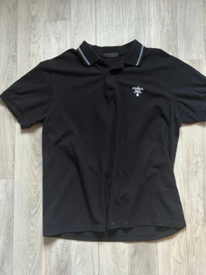 Svart pikétröja från Prada - Svart pikétröja från Prada med klassisk krage och två knappar framtill. Tröjan har diskret logga på bröstet och vita ränder på kragen. Perfekt för en stilren och avslappnad look.