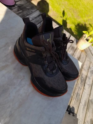 Bruna sneakers från Michael Kors - Snygga bruna sneakers från Michael Kors med svarta och mörkbruna detaljer samt orange sula. Skorna har snörning, högre skaft och tydlig MK-logga på plösen. Materialmix av textil och syntet ger en modern look.