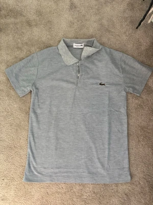 Ljusgrå pikétröja från Lacoste - Snygg ljusgrå pikétröja från Lacoste med klassisk krage och knappar framtill. Tröjan har korta ärmar och den ikoniska krokodilloggan broderad på bröstet. Perfekt för en clean och stilren look.