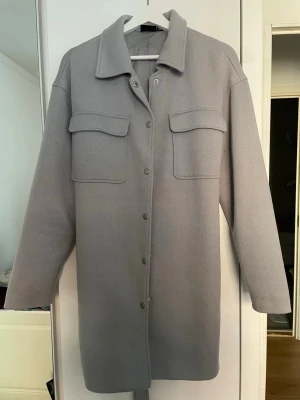 Ljusgrå kappa med fickor - Snygg ljusgrå kappa i längre modell med raka ärmar och två stora bröstfickor med lock. Stängs med tryckknappar framtill och har klassisk krage. Perfekt lager-på-lager-plagg i ett mjukt, tjockare material.