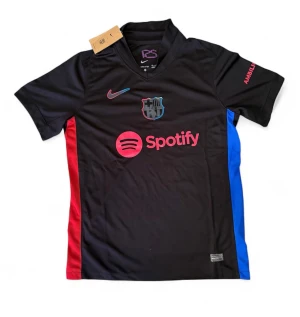 Svart FC Barcelona matchtröja Nike - Snygg svart FC Barcelona matchtröja från Nike med röda och blå detaljer på sidorna. Tröjan har klubbmärke, Nike-logga och Spotify-tryck på bröstet samt UNHCR-logga på ryggen. Tillverkad i lätt och ventilerande material, perfekt för fotbollsträning.
