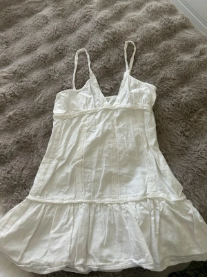 Vit sommarklänning från Pull&Bear - Söt vit kortklänning från Pull&Bear med tunna axelband och v-ringning. Klänningen har volang nedtill och är tillverkad i ett lätt bomullstyg, perfekt för varma dagar. Enkel och fräsch design som passar till många tillfällen.