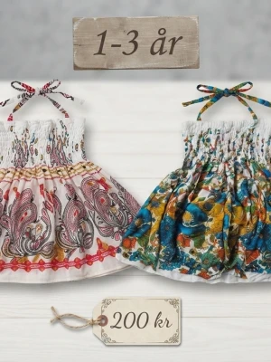 Färgglada sommarklänningar 1-3 år - Två söta korta klänningar för barn 1-3 år med smock upptill och knytband i nacken. Ena klänningen är vit med paisley- och blommönster i rött och rosa, den andra har blått och gult blommönster. Perfekta för sommardagar!