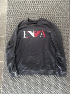 Svart sweatshirt från Emporio Armani - Snygg svart sweatshirt från Emporio Armani med stort EVA-logotyptryck i vitt och rött på bröstet. Tröjan har rund halsringning, långa ärmar och ribbade muddar vid ärmslut och nederkant. Perfekt för en avslappnad och trendig look.