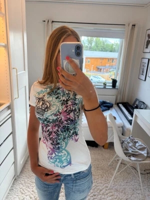Vit t-shirt med färgglatt tryck - Säljer en vit t-shirt med korta ärmar och ett färgglatt tryck i på framsidan. T-shirten sitter tajt, men har ett otroligt mjukt material. En snygg t-shirt med ett intryck av 2000-tal och passar perfekt till ett par stilrena jeans. 