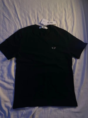 CDG T-shirt - Svart & svart hjärta - Storlek S - Snygg svart t-shirt med ett broderat hjärta med ögon på bröstet. Klassisk rund hals och korta ärmar. Perfekt basplagg med unik detalj som sticker ut i mängden.