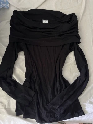 Svart offshoulder topp med lång ärm - Trendig svart offshoulder topp med långa ärmar och bred krage som vilar över axlarna. Toppens material känns mjukt och följsamt, perfekt för en stilren look. Passformen är slim och toppen är enkel att matcha med olika outfits.