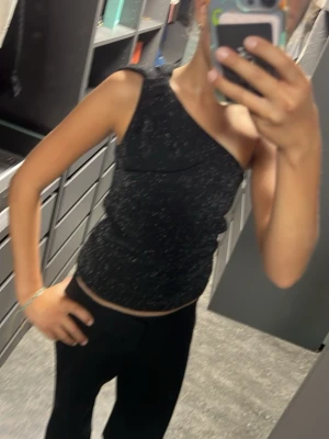 Svart glittrig oneshoulder topp - Snygg svart oneshoulder topp med glittrigt tyg och en liten rosettdetalj vid axeln. Toppen är tight och croppad, perfekt för dig som vill sticka ut lite extra. Passar till både jeans och kjol för en trendig look.❤️