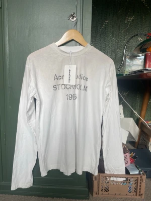 Acne studios longsleeve - Vit långärmad topp från Acne Studios med tryck 'Acne Studios STOCKHOLM 1996' framtill. Tillverkad i mjuk bomull och har en enkel, clean design med normal passform. Perfekt till jeans eller kjol för en stilren look.