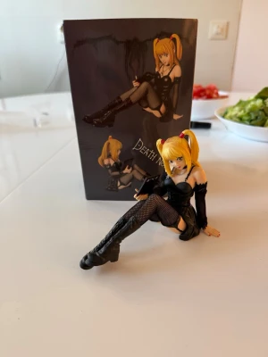 Death Note Misa Amane figur - Säljer en detaljerad Misa Amane-figur från Death Note. Hon har blont hår i tofsar, svart klänning, nätstrumpor och höga stövlar. Figuren håller en svart bok och har gotisk stil. Perfekt för samling!