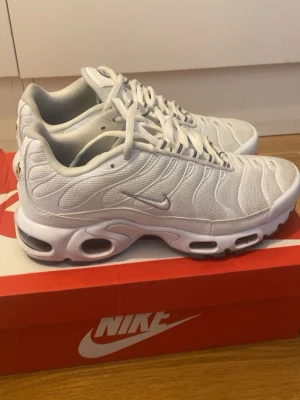 Vita Nike Air Max Plus sneakers - Säljer ett par vita Nike Air Max Plus sneakers med mesh- och syntetmaterial. Skorna har den klassiska TN-designen med vågiga linjer, synlig Air-dämpning i sulan och små silverdetaljer. Perfekta för dig som gillar sportig och clean stil. De är helt nya använda ett par gånger säljer de för de är liten på mig och pris kan diskuteras 
