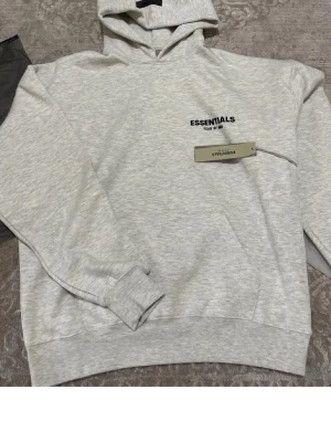 Ljusgrå Essentials hoodie med huva - Snygg ljusgrå hoodie från Essentials med svart logga på bröstet. Klassisk modell med huva och ribbade muddar vid ärmar och nederkant. Tillverkad i mjukt material som känns skönt mot huden. Perfekt för en avslappnad och trendig look.