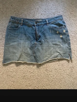 Blå jeanskjol - Säljer en blå jeanskjol i kort modell med rå, fransig kant nertill och slitna detaljer framtill. Kjolen har klassiska fickor både fram och bak samt bälteshällor. Perfekt för en avslappnad och trendig look.