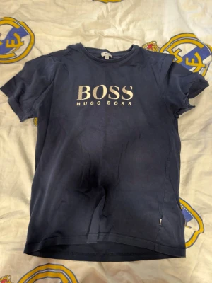 Blå Hugo Boss t-shirt med tryck - Snygg mörkblå t-shirt från Hugo Boss med stort vitt logotryck på bröstet. Klassisk rund hals och korta ärmar. Perfekt för en stilren och avslappnad look.