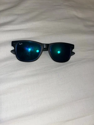 Svarta Ray-Ban solglasögon Wayfarer - Snygga svarta Ray-Ban Wayfarer solglasögon med klassisk form och mörka blåtonade spegelglas. Perfekta för att höja din stil och skydda ögonen mot solen. Diskret Ray-Ban-logga på bågen och glaset.