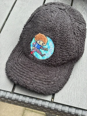 Svart teddykeps med brodyr - Säljer en svart keps i teddy-material med en färgglad brodyr framtill som föreställer en tecknad figur och texten 'Boylan' på blå bakgrund. Kepsen har en mjuk och fluffig struktur som ger en unik look.