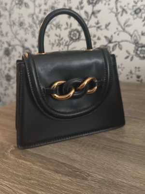 Svart handväska med gulddetaljer - Stilren svart handväska i slätt konstläder med guldfärgade kedjedetaljer på locket. Väskan har ett topphandtag och magnetlås. Insidan har flera fack för enkel organisering. Perfekt för dig som gillar trendiga accessoarer med en lyxig touch.
