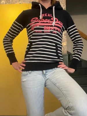 Randig hoodie från Superdry - Snygg svart hoodie från Superdry med vita horisontella ränder och stor rosa logga på bröstet. Hoodien har vit dragsko i huvan och en klassisk känguruficka framtill. Perfekt för en avslappnad streetstyle-look.