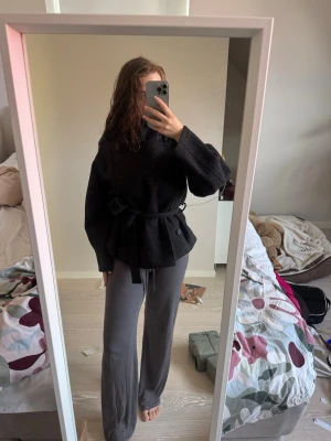 Svart kappa med knytskärp - Snygg svart kappa med breda ärmar och knytskärp i midjan. Kappan har en avslappnad passform och hög krage, perfekt för kyligare dagar. Materialet ser ut att vara ullblandning och modellen är trendigt oversized.