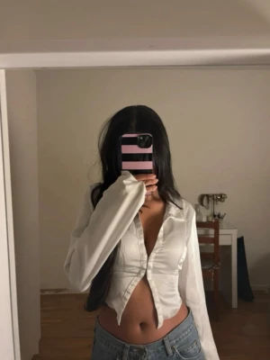 Vit croppad blus med v-ringning - Säljer en vit croppad blus med v-ringning och långa ärmar. Toppen har en snygg omlott-look framtill och är lätt figursydd för en smickrande passform. Perfekt att styla med jeans eller kjol för en trendig look.