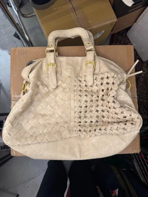 Beige handväska med flätad detalj - Stilren beige handväska i mjukt material med dubbla handtag och guldfärgade metalldetaljer. Framsidan har en unik flätad design på ena halvan och slät yta på andra. Väskan har dragkedja upptill och en ficka med dragkedja på baksidan. Perfekt för dig som gillar trendiga accessoarer. Finns att hämta i Gröndal.