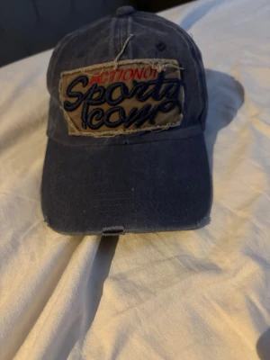 Blå keps med broderad patch - Snygg blå keps med böjd skärm och en stor beige patch framtill med texten 'Action No1 Sports Icon' i rött och blått. Kepsen har en vintage look med slitna detaljer och är troligen gjord i bomullstyg. Perfekt för en avslappnad streetwear-stil.