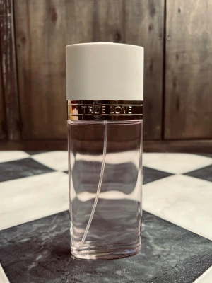 True Love parfym från Elizabeth Arden 100ml - True Love parfym från Elizabeth Arden med en elegant cylinderformad flaska i transparent glas och subtil rosa nyans. Flaskan har en vit matt kork och ett guldfärgat band med texten 'TRUE LOVE' runt halsen. Perfekt för dig som gillar stilrena och klassiska dofter.