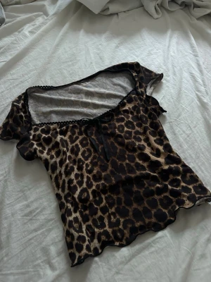 Leopardmönstrad topp med rosett - Snygg leopardmönstrad topp med en liten svart rosett framtill. Toppen har vågig kant nertill och spetsdetalj vid urringningen. Perfekt för dig som vill sticka ut lite extra.